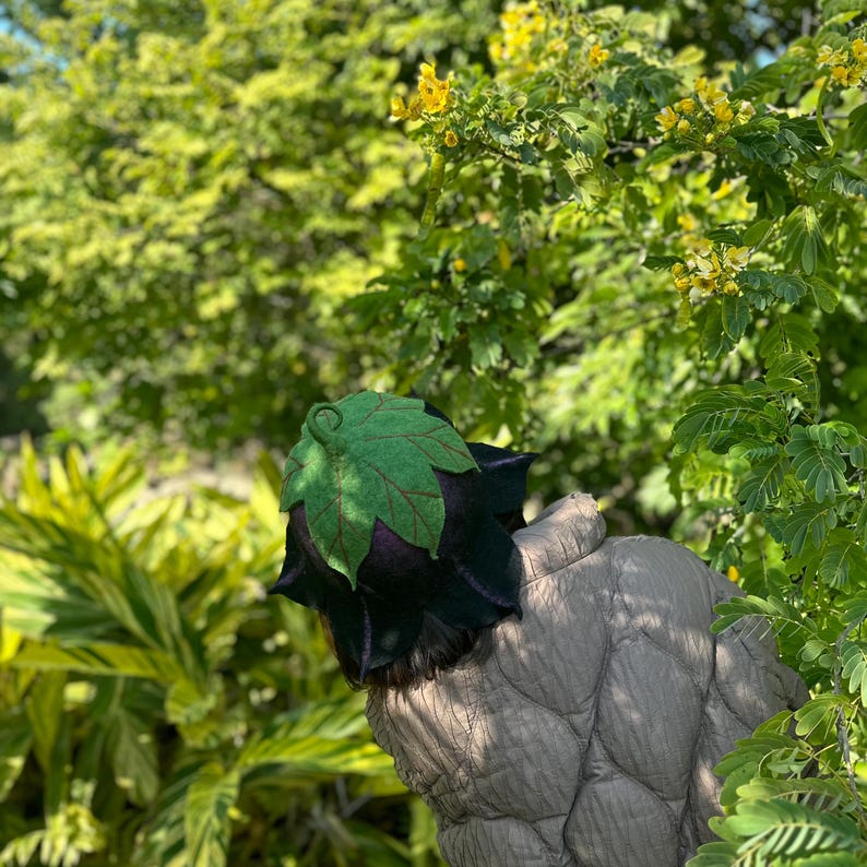 Puede incluir: Un sombrero &uacute;nico con un dise&ntilde;o de hoja. El sombrero presenta una hoja verde en la parte superior y una base de color p&uacute;rpura oscuro y negro. El sombrero est&aacute; rodeado de follaje verde y flores amarillas.