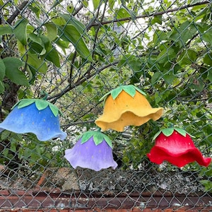 Peut inclure: Quatre chapeaux en feutre en forme de fleurs, bleus, jaunes, violets et rouges, chacun avec des accents de feuilles vertes. Les chapeaux sont accrochés à une clôture en treillis métallique, avec un mur de briques et de la verdure en arrière-plan.