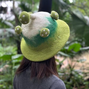 Könnte beinhalten: Ein grüner und weißer Filzhut mit Pilz-förmigen Verzierungen. Der Hut hat eine breite Krempe und eine abgerundete Krone.