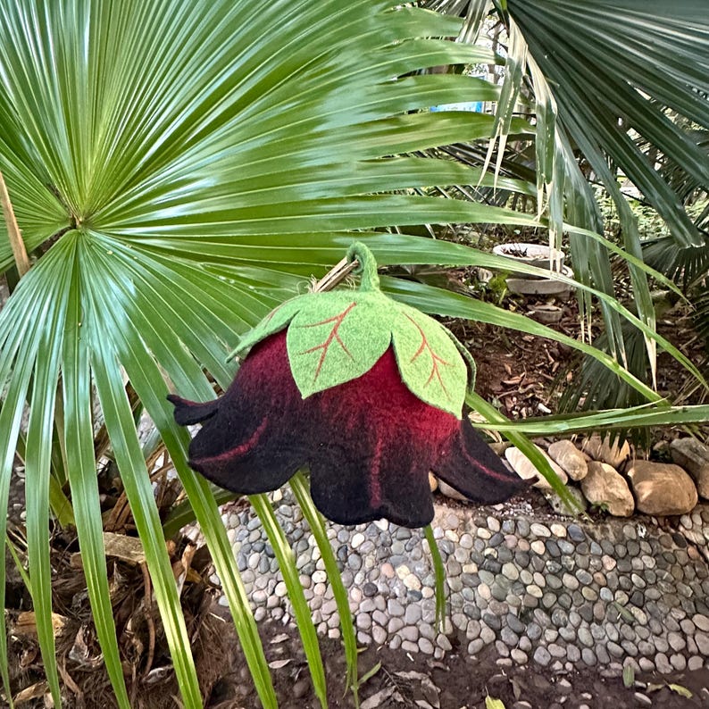 Puede incluir: Una flor de fieltro hecha a mano en tonos negro y burdeos, con hojas verdes y venas rojas. La flor est&aacute; suspendida frente a un fondo de hojas de palmera verdes y un camino de piedra, creando una exhibici&oacute;n natural y art&iacute;stica.