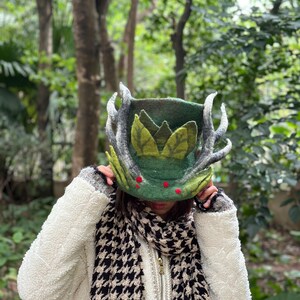 May include: A green felt hat with antlers and leaves, worn by a person with a black and white scarf.
