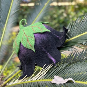 Puede incluir: Un sombrero caprichoso, hecho a mano, con forma de berenjena. El sombrero es principalmente de color p&uacute;rpura oscuro y negro, con una hoja y un tallo verdes. La hoja tiene vetas rojas. El sombrero est&aacute; hecho de fieltro.