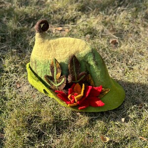 May include: A green felt hat with a pointed crown and a curled brim. The hat is decorated with felt leaves and a red felt flower.
