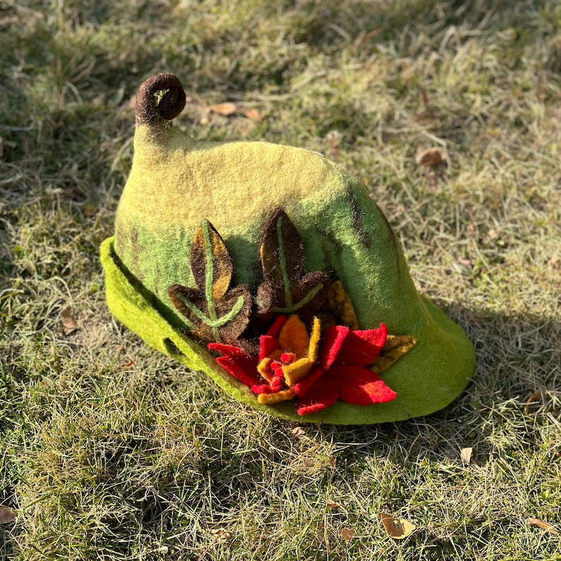 May include: A green felt hat with a pointed crown and a curled brim. The hat is decorated with felt leaves and a red felt flower.