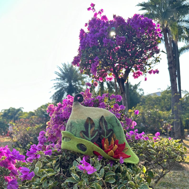 May include: A green felt hat with a pointed crown and a curled brim. The hat is decorated with felt leaves and flowers. The hat is sitting on a bed of purple flowers.