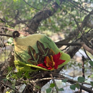 May include: A green felt hat with a leaf design and a red flower accent. The hat is sitting on a branch.