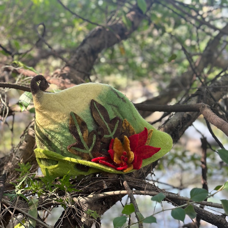 May include: A green felt hat with a leaf design and a red flower accent. The hat is sitting on a branch.