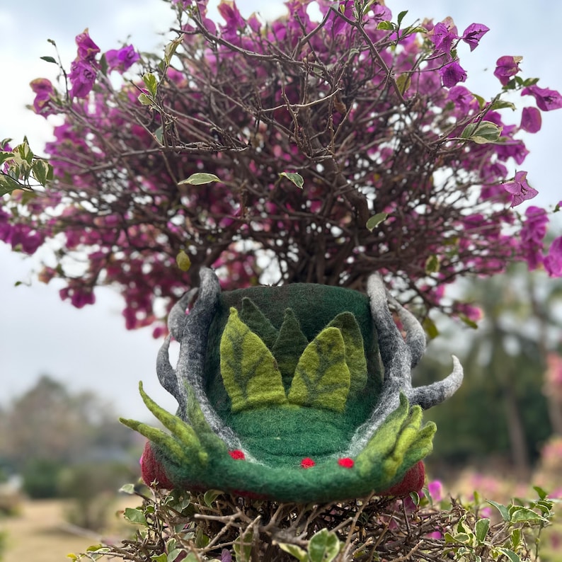 May include: A green felt hat with a leafy design and antlers. The hat is sitting on a branch of a bush with pink flowers.