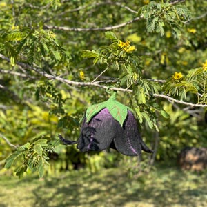 Puede incluir: Un sombrero artesanal en forma de flor, de fieltro negro y morado, con detalles de hojas verdes. El sombrero est&aacute; suspendido de una rama de &aacute;rbol, mostrando su dise&ntilde;o &uacute;nico y artesan&iacute;a. Un accesorio caprichoso.