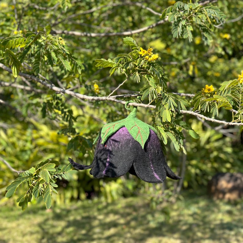 Puede incluir: Un sombrero artesanal en forma de flor, de fieltro negro y morado, con detalles de hojas verdes. El sombrero est&aacute; suspendido de una rama de &aacute;rbol, mostrando su dise&ntilde;o &uacute;nico y artesan&iacute;a. Un accesorio caprichoso.