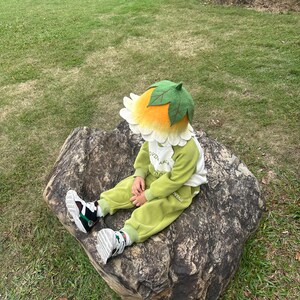 May include: A child wearing a flower-shaped hat with a green leaf top and yellow petals. The child is also wearing a green and white outfit and sitting on a large rock. The outfit includes the text "OVIN AM".