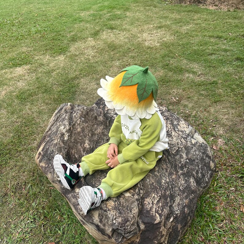 May include: A child wearing a flower-shaped hat with a green leaf top and yellow petals. The child is also wearing a green and white outfit and sitting on a large rock. The outfit includes the text "OVIN AM".