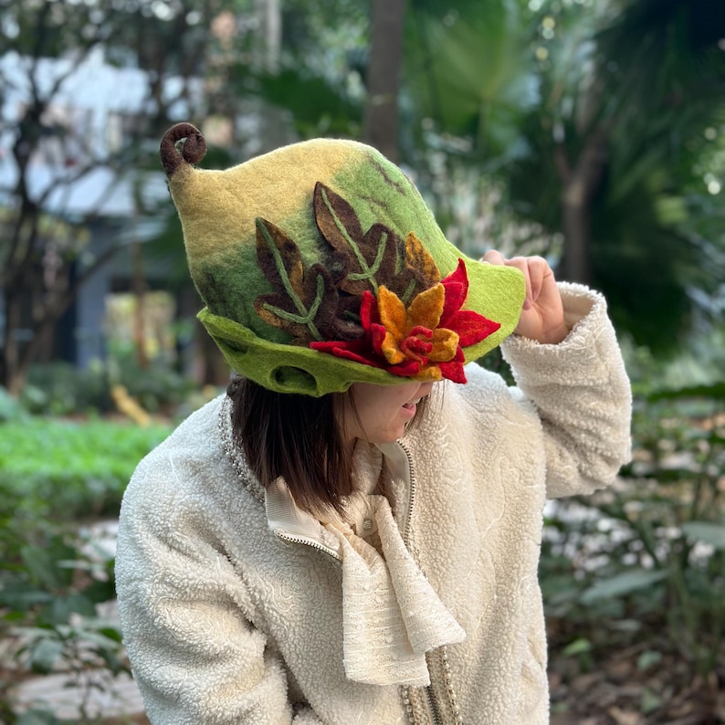 May include: A green, yellow, and brown felt hat with a flower and leaves. The hat has a pointed top and a curled brim.