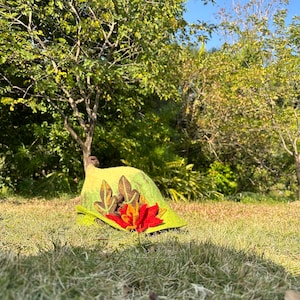 May include: A green felt hat with a red flower and leaf embellishments. The hat is lying on a bed of green grass.