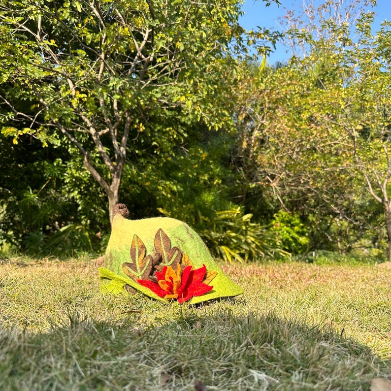 May include: A green felt hat with a red flower and leaf embellishments. The hat is lying on a bed of green grass.