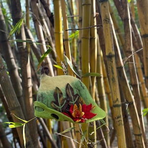 May include: A green and yellow felt hat with a flower and leaves on the side. The hat is sitting in a bamboo forest.