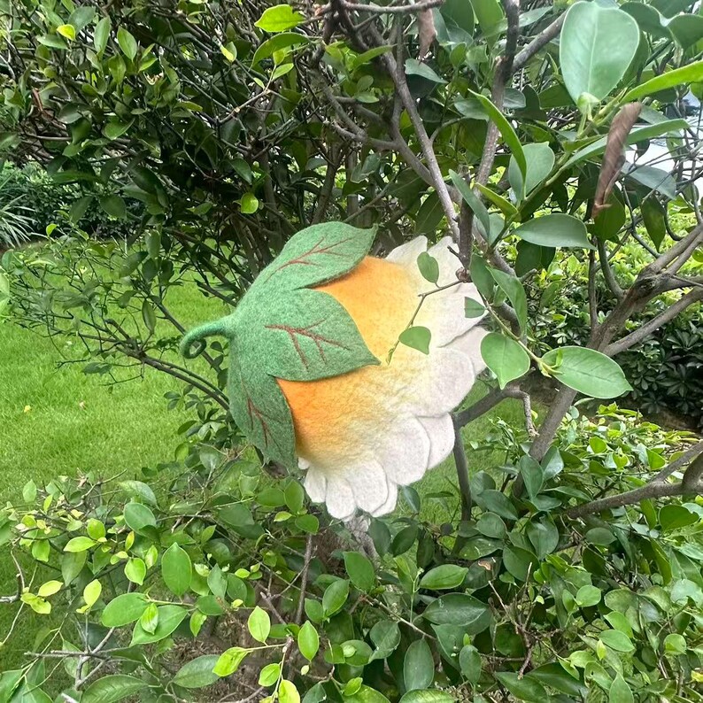 May include: A felt flower decoration with white petals, a yellow center, and green leaves. The handmade floral accent is set against a backdrop of green foliage.