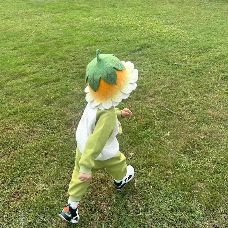 May include: A child wearing a sunflower-themed costume, including a hat and a green and white outfit, walks on a grassy lawn. The hat features a yellow center, white petals, and green leaves. The outfit is green with white accents.