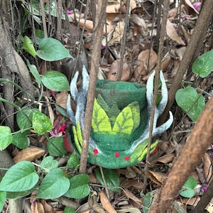 May include: A green felt hat with a crown of leaves and antlers. The hat has red accents and is surrounded by green foliage.