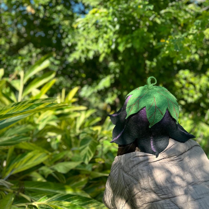 Puede incluir: Un sombrero caprichoso con un dise&ntilde;o &uacute;nico, con una base en forma de campana de color p&uacute;rpura oscuro y una parte superior en forma de hoja verde. El dise&ntilde;o del sombrero incluye detalles de venas rojas en las hojas. El sombrero se encuentra sobre un fondo de exuberante follaje verde.