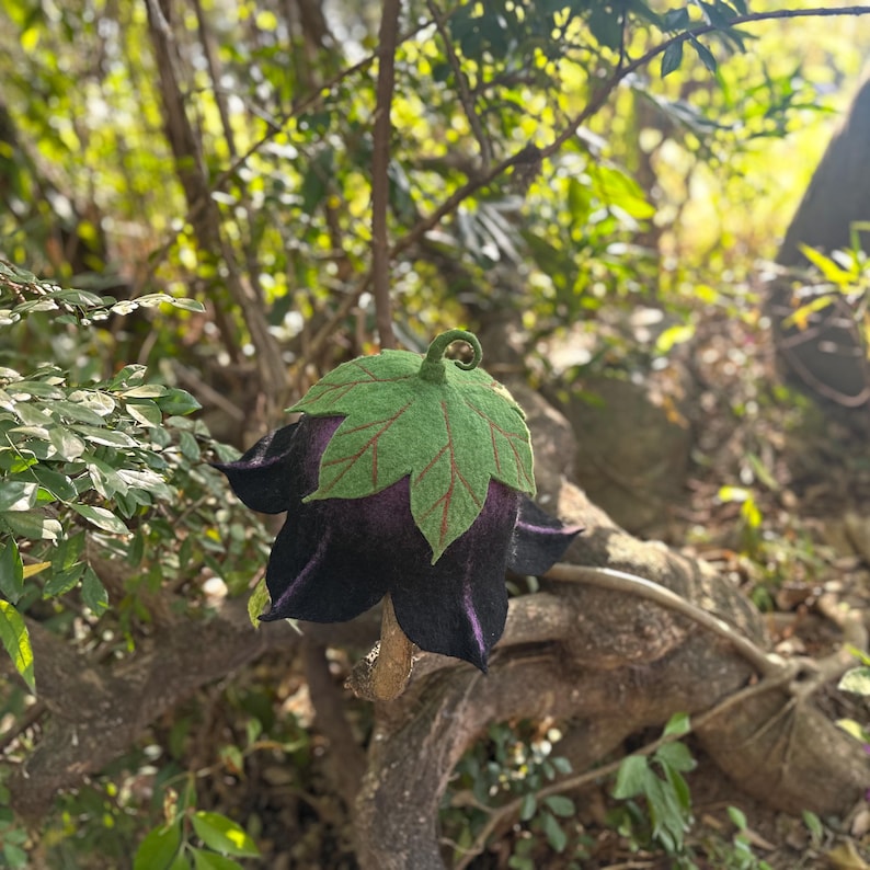 Puede incluir: Una flor de fieltro hecha a mano con una flor de color p&uacute;rpura oscuro y una parte superior en forma de hoja verde. La flor de fieltro est&aacute; unida a una rama natural, sobre un fondo borroso de follaje verde y ramas de &aacute;rboles.