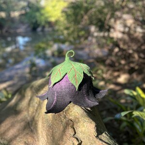 Puede incluir: Un sombrero de fieltro hecho a mano con forma de campanilla. El sombrero presenta una campana de color p&uacute;rpura oscuro con bordes negros, rematada con hojas verdes con vetas rojas. El sombrero est&aacute; sobre un fondo natural borroso.