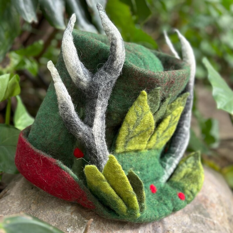 May include: A green felt hat with a red brim, decorated with white and gray felt antlers, green felt leaves, and red felt berries.