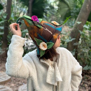Puede incluir: Un sombrero de fieltro artesanal con forma puntiaguda, similar a un sombrero de elfo. El sombrero presenta una base marrón con detalles en azul, naranja y blanco. Está adornado con hojas de fieltro verdes y flores de tela coloridas en tonos rosa y crema. Un accesorio único.