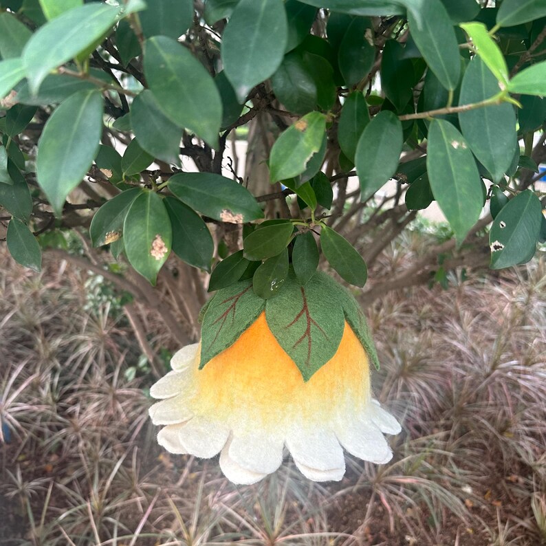 May include: A handmade felt flower ornament with a white petal design and a yellow center. The ornament is adorned with green felt leaves with red veins. The flower is hanging from a tree branch, creating a whimsical and natural aesthetic.