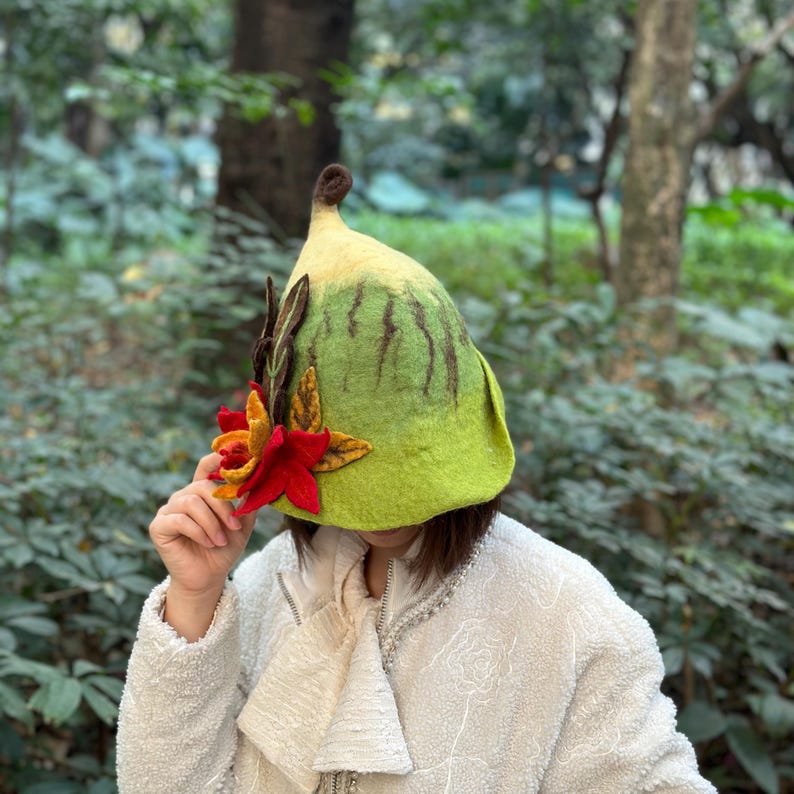 May include: A green and yellow felt hat with a flower and leaves on the side. The hat is shaped like a bell and has a pointed top.