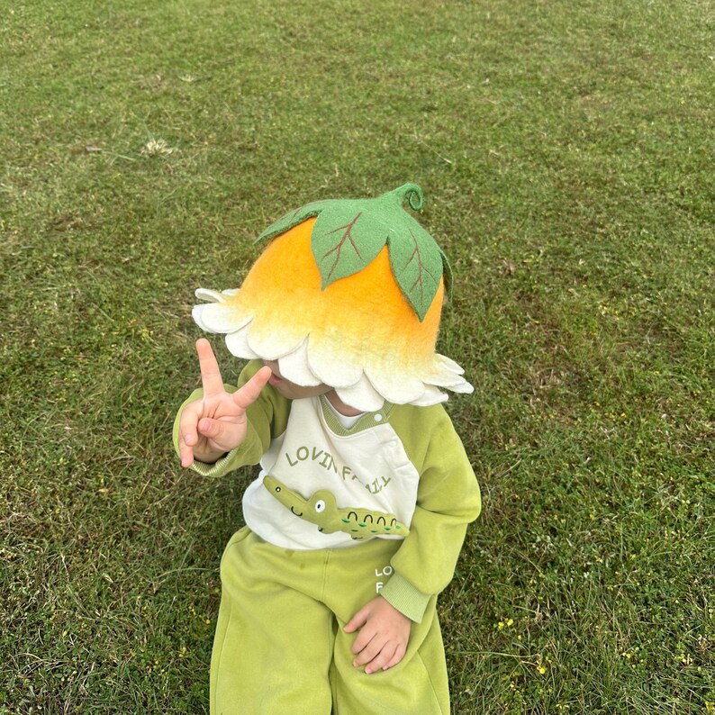 May include: A child wearing a whimsical flower hat with a yellow and white petal design and green leaves. The child is also wearing a green outfit with a crocodile graphic and the text "LOVIN' FALL". The child is lying on green grass.