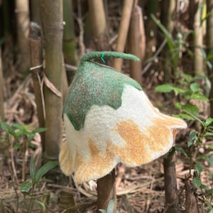 May include: A handmade felt hat in the shape of a flower. The hat features a white and yellow petal design with a green top and stem. The hat is set against a natural background of bamboo stalks and foliage.