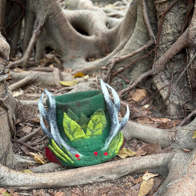 May include: A green felt hat with antlers and green leaves. The hat is sitting on a tree root.