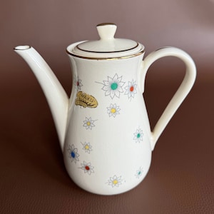 Op de afbeelding: Witte theepot van keramiek met een gouden rand en een bloemenpatroon. De theepot heeft een deksel en een handvat. Het bloemenpatroon heeft witte bloemen met gele, rode, blauwe en groene centra. De theepot heeft een gouden label met de tekst "Waku Coffee".