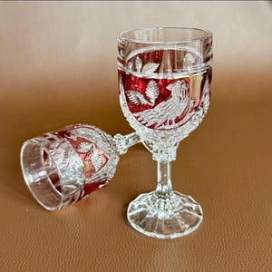 Könnte beinhalten: Zwei klare Kristall-Weingläser mit roter Innenseite und Vogel- und Blattdesign. Ein Glas steht aufrecht, das andere liegt auf der Seite. Die Gläser haben ein detailliertes Schliffmuster und einen Stiel.