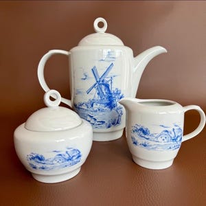 Puede incluir: Juego de té de porcelana blanca con diseños azules de molinos de viento y paisajes. El juego incluye una tetera, un azucarero con tapa y una jarra para crema. La tetera tiene un asa curva y un pico vertedor.