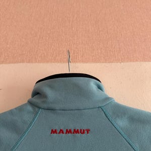 Puede incluir: Chaqueta polar azul claro con cuello alto y la palabra "MAMMUT" bordada en rojo en la espalda. La chaqueta est&aacute; colgada de una percha met&aacute;lica.