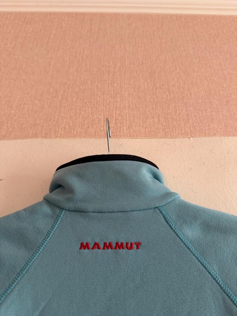Puede incluir: Chaqueta polar azul claro con cuello alto y la palabra "MAMMUT" bordada en rojo en la espalda. La chaqueta est&aacute; colgada de una percha met&aacute;lica.