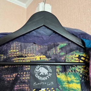 K&ouml;nnte beinhalten: Eine dunkelblaue Jacke mit mehrfarbigem Innenfutter mit abstrakten Mustern in Gelb, Gr&uuml;n und Violett. Innen ist ein schwarzes quadratisches Etikett mit einem wei&szlig;en Mammut-Logo, dem Text Mammut Garantie und Mountain Life aufgen&auml;ht.