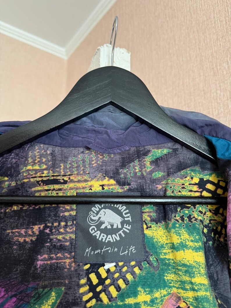 K&ouml;nnte beinhalten: Eine dunkelblaue Jacke mit mehrfarbigem Innenfutter mit abstrakten Mustern in Gelb, Gr&uuml;n und Violett. Innen ist ein schwarzes quadratisches Etikett mit einem wei&szlig;en Mammut-Logo, dem Text Mammut Garantie und Mountain Life aufgen&auml;ht.
