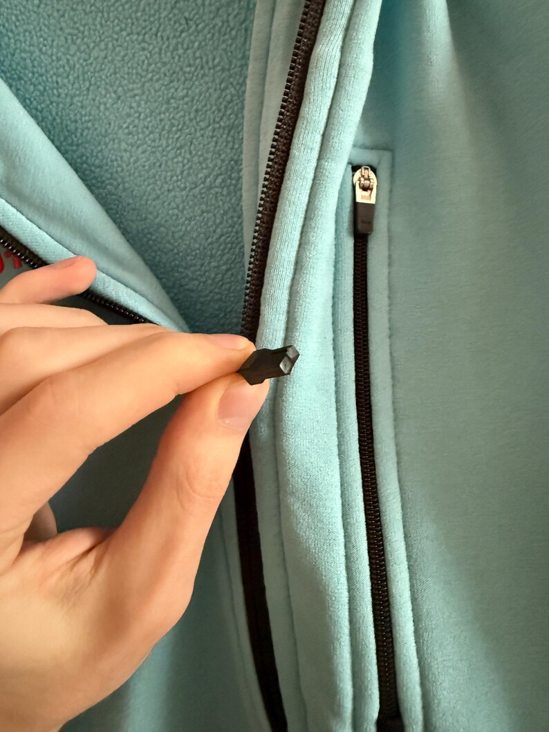 Puede incluir: Primer plano de una chaqueta de forro polar azul claro con una cremallera negra. La cremallera est&aacute; parcialmente abierta y una mano sostiene el tirador. La chaqueta tiene un aspecto suave y texturizado.