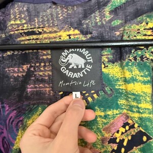K&ouml;nnte beinhalten: Eine Jacke mit einem mehrfarbigen abstrakten Muster in Gr&uuml;n-, Gelb-, Lila- und Schwarzt&ouml;nen. Ein schwarzes Etikett zeigt ein wei&szlig;es Mammut-Logo, den Text Mammut Garantie und die Worte Mountain Life. Ein kleines wei&szlig;es Etikett zeigt die Gr&ouml;&szlig;e L.