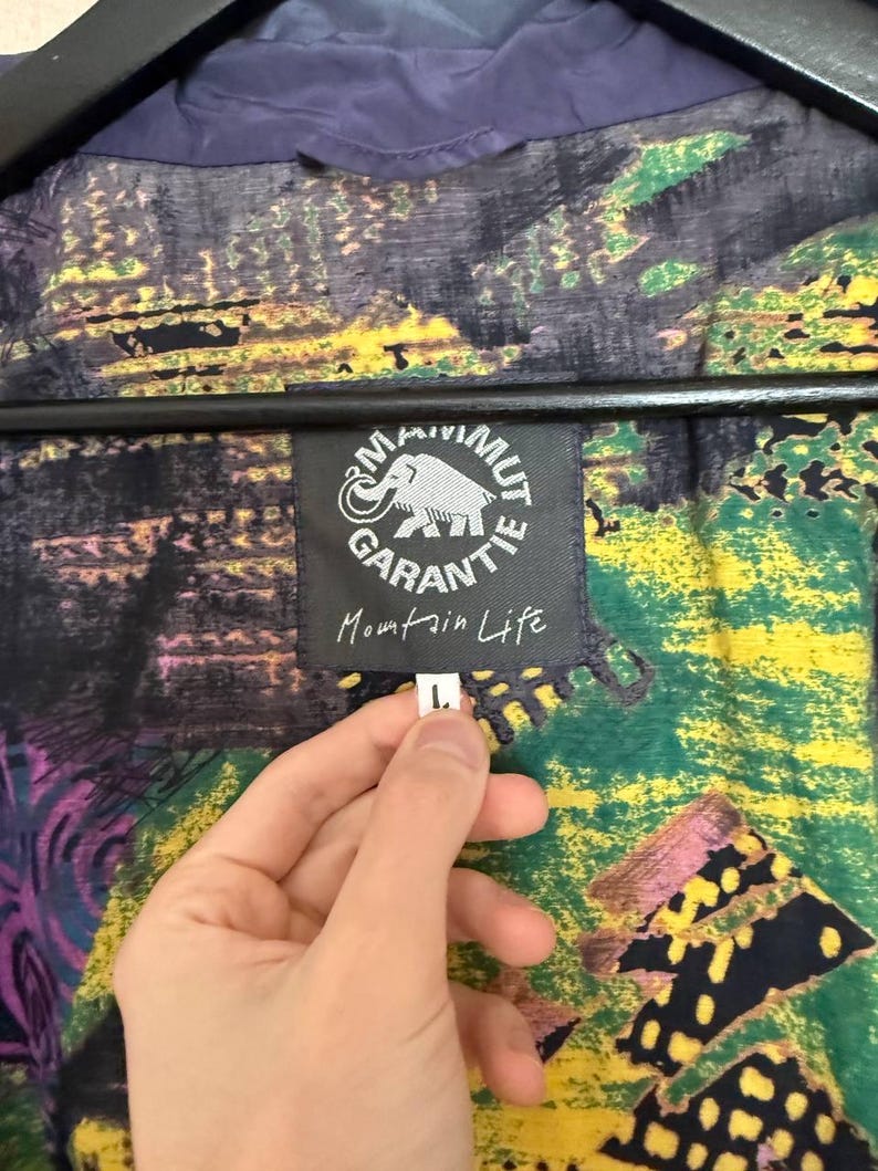 K&ouml;nnte beinhalten: Eine Jacke mit einem mehrfarbigen abstrakten Muster in Gr&uuml;n-, Gelb-, Lila- und Schwarzt&ouml;nen. Ein schwarzes Etikett zeigt ein wei&szlig;es Mammut-Logo, den Text Mammut Garantie und die Worte Mountain Life. Ein kleines wei&szlig;es Etikett zeigt die Gr&ouml;&szlig;e L.