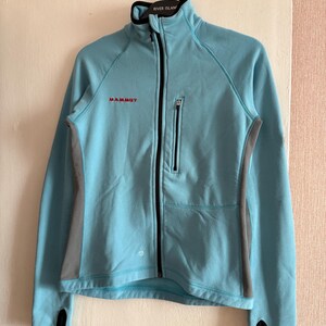 Puede incluir: Chaqueta Mammut azul claro con cremallera completa, ribetes negros y paneles laterales grises. La chaqueta tiene un cuello alto y un peque&ntilde;o bolsillo con cremallera en el pecho. Los pu&ntilde;os tienen orificios para los pulgares.