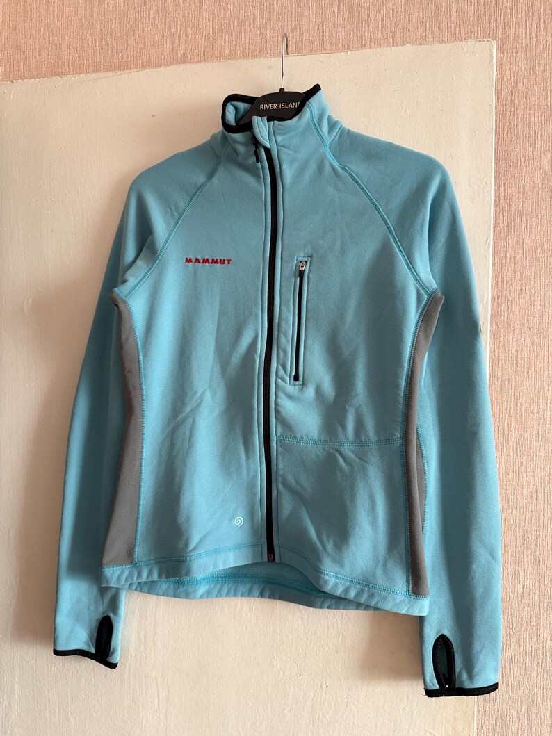 Puede incluir: Chaqueta Mammut azul claro con cremallera completa, ribetes negros y paneles laterales grises. La chaqueta tiene un cuello alto y un peque&ntilde;o bolsillo con cremallera en el pecho. Los pu&ntilde;os tienen orificios para los pulgares.
