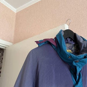 K&ouml;nnte beinhalten: Eine farblich abgesetzte Jacke mit Paneelen in ged&auml;mpftem Lila und Petrol. Die Jacke verf&uuml;gt &uuml;ber einen hohen Kragen, eine abnehmbare Kapuze mit lila und petrolfarbenen Akzenten sowie eine Frontleiste mit sichtbaren Druckkn&ouml;pfen.