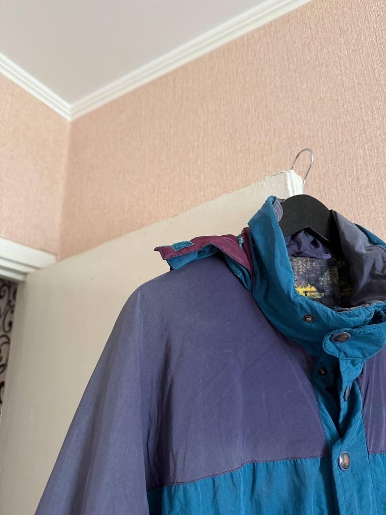 K&ouml;nnte beinhalten: Eine farblich abgesetzte Jacke mit Paneelen in ged&auml;mpftem Lila und Petrol. Die Jacke verf&uuml;gt &uuml;ber einen hohen Kragen, eine abnehmbare Kapuze mit lila und petrolfarbenen Akzenten sowie eine Frontleiste mit sichtbaren Druckkn&ouml;pfen.