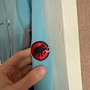 Puede incluir: Chaqueta azul claro con un panel gris y un parche con el logotipo negro y rojo en la manga. El logotipo presenta un elefante negro con un colmillo blanco dentro de un c&iacute;rculo rojo. La chaqueta tiene una cremallera.