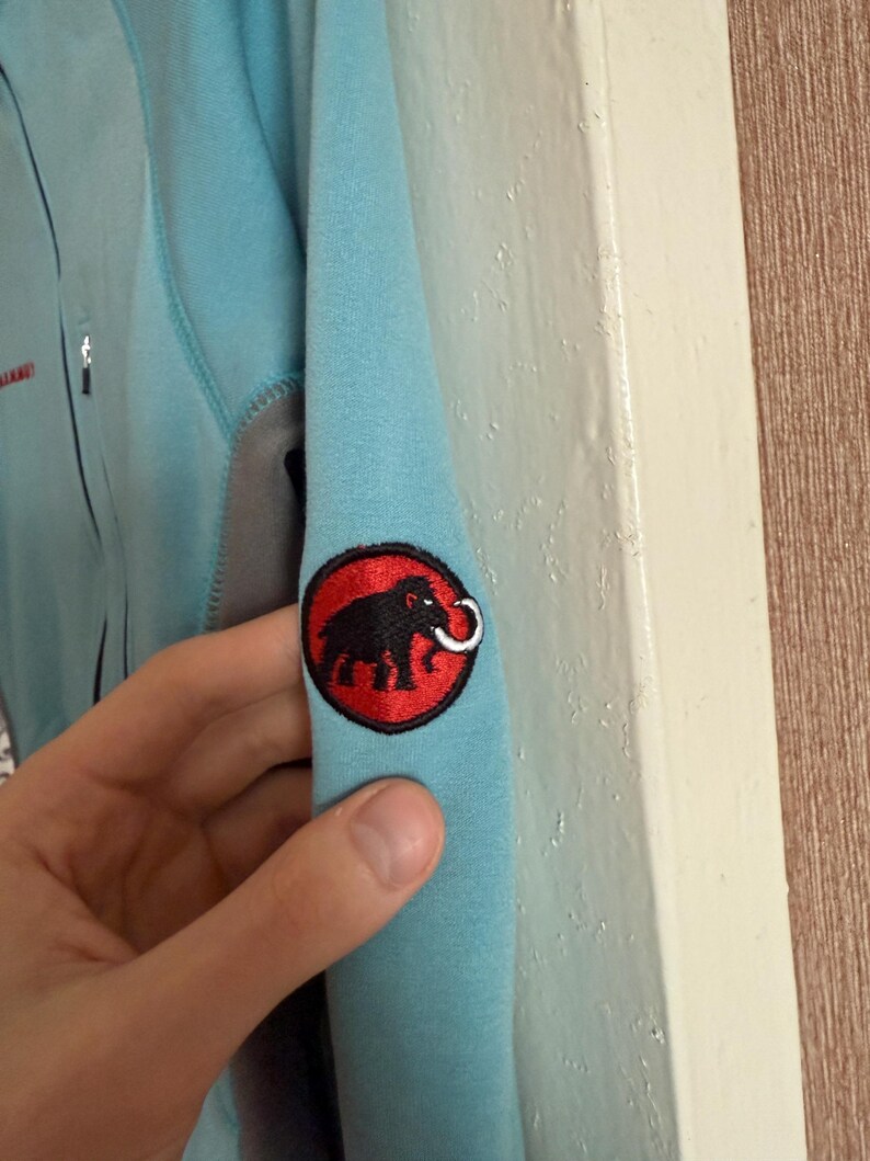Puede incluir: Chaqueta azul claro con un panel gris y un parche con el logotipo negro y rojo en la manga. El logotipo presenta un elefante negro con un colmillo blanco dentro de un c&iacute;rculo rojo. La chaqueta tiene una cremallera.