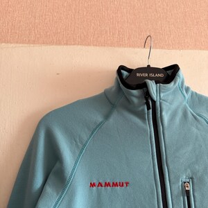 Puede incluir: Chaqueta polar azul claro con cremallera negra y un peque&ntilde;o bolsillo con cremallera. La chaqueta tiene la palabra "MAMMUT" bordada en rojo en el pecho. El cuello es negro y la chaqueta est&aacute; colgada en una percha con "RIVER ISLAND".